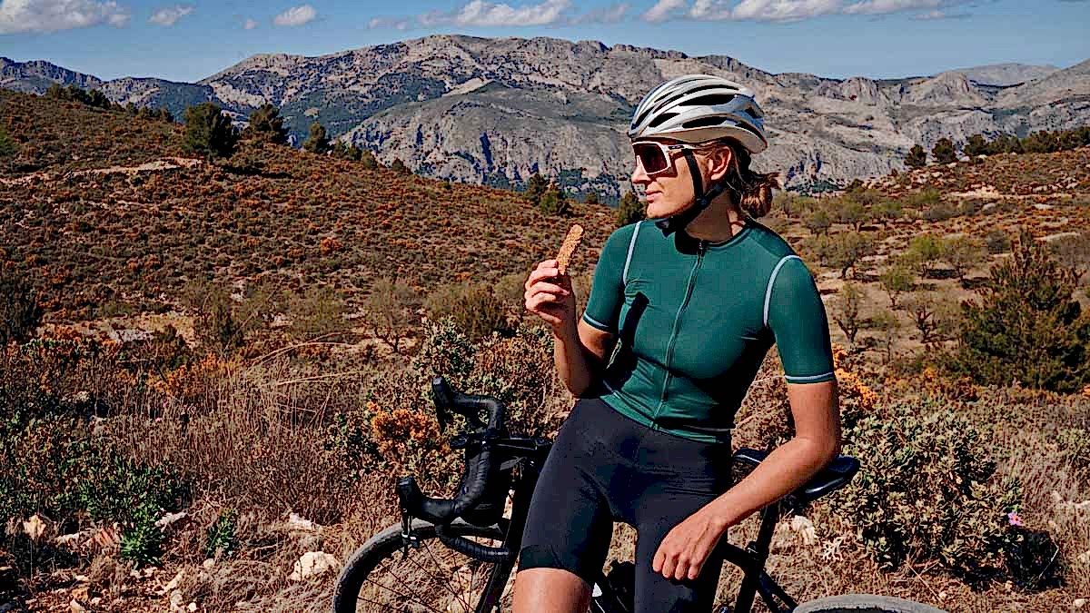 Female Cyclist Wearing Helmet And Sunglasses Resting On Her Bike And Eating Nutrition Bar With Mountains In The Background
