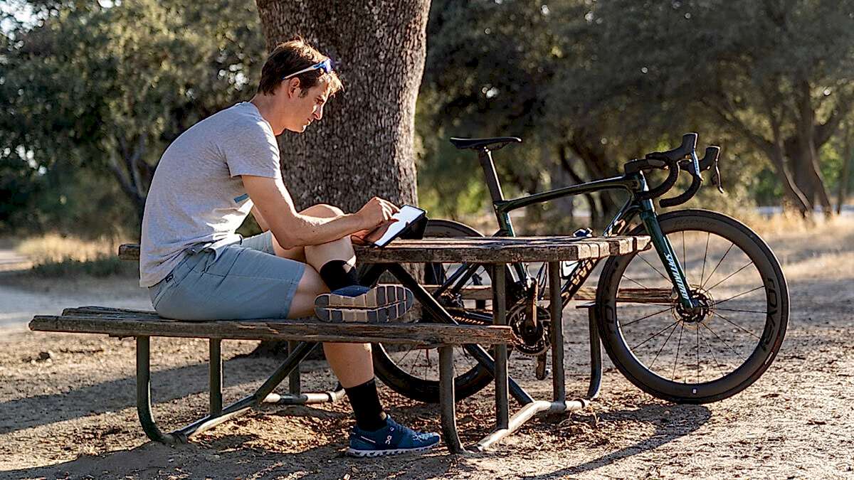 Running Cycling Coach Sitting On Laptop At The Park On Picnic Table Next To Back Setting Prices For Coaching Services